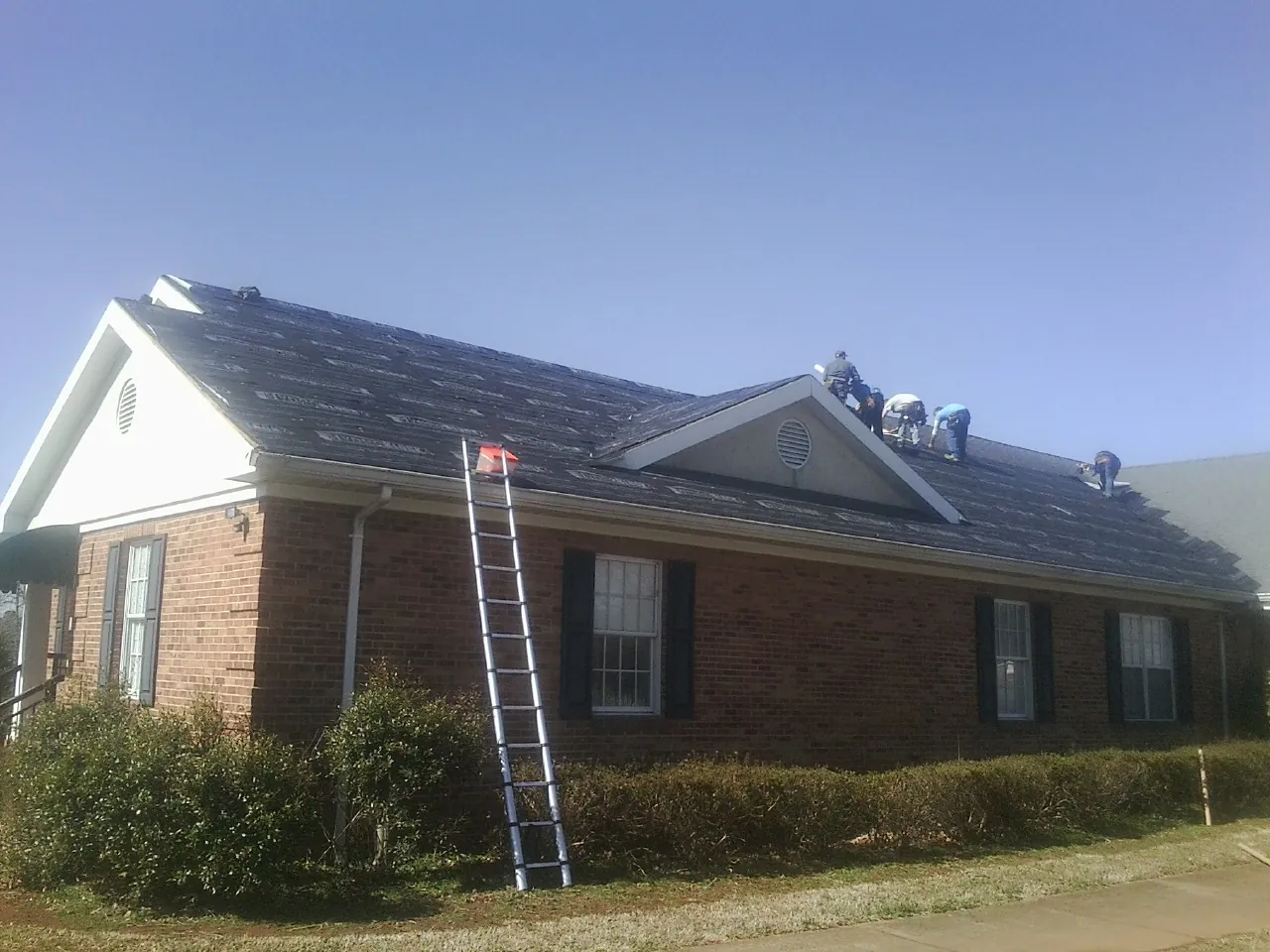 Crew tearing shingles off a brick ranch roof with a ladder set — Emergency Roof Repair work by our crew in Winger