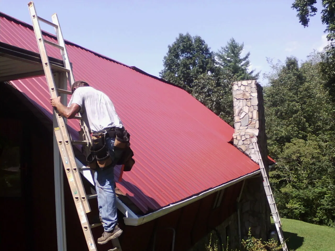 Roofer on a ladder beside a red metal roof and stone chimney — Soffit & Fascia Repair by Roofing Brothers in Winger
